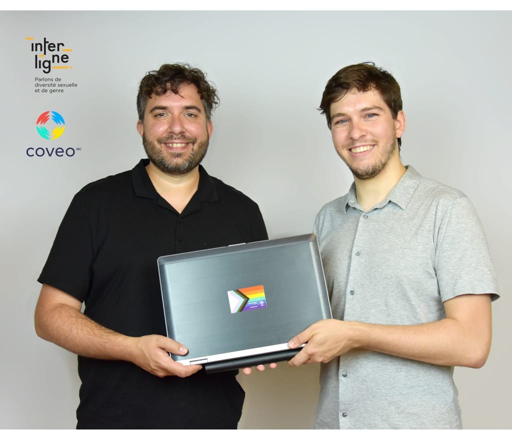 A photo of two men holding a laptop that has an LBGTQ+ sticker on its lid.