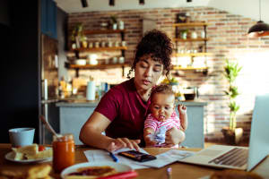 Multiracial woman with child, writing on a notepad in front of her laptop